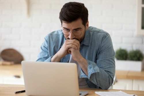 Un hombre con gafas y camisa de mezclilla mirando fijamente la pantalla de su laptop con expresión pensativa, concentrada o preocupada, apoyando sus manos juntas cerca de la barbilla