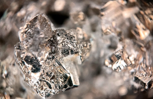 Fotografía macro de un mineral cristalino brillante con facetas geométricas plateadas o doradas claras, similar a la pirita, mostrando texturas rugosas y reflejos metálicos.