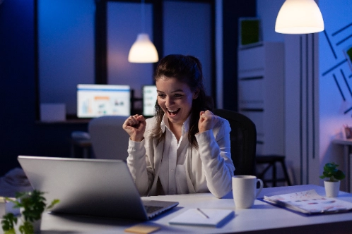 una mujer sonríe y celebra con los puños cerrados frente a su laptop en una oficina iluminada con tonos azules nocturnos.