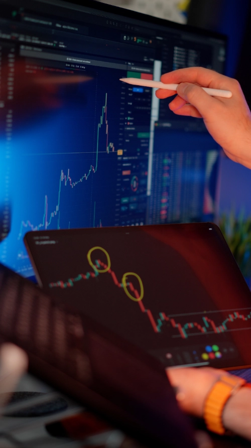Una mano apunta con un lápiz óptico hacia un gran monitor con gráficos financieros, mientras otra pantalla en primer plano muestra una tendencia a la baja con zonas marcadas en círculos amarillos.