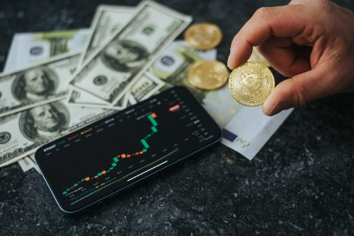 Una mano sostiene una moneda física de Bitcoin sobre un teléfono móvil con gráficos financieros y billetes de dólar.