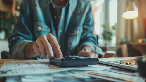 Una persona vestida con una chaqueta de mezclilla utiliza una calculadora sobre un escritorio lleno de papeles y documentos, junto a una computadora portátil.