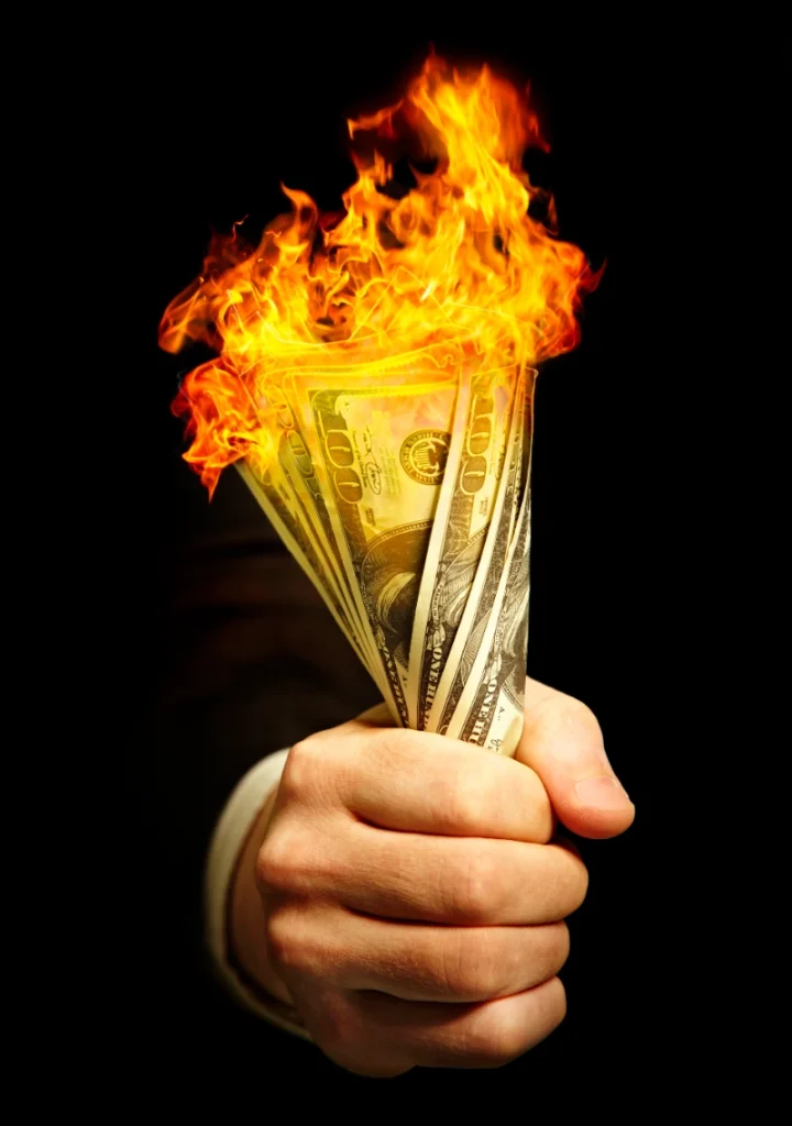 Un puño sostiene con fuerza un mazo de billetes de cien dólares ardiendo en llamas sobre un fondo negro.
